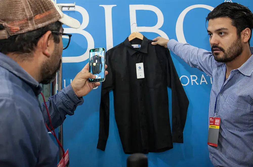 Empresario mostrando una prenda a un visitante que la fotografía con su celular, ejemplo de estrategias digitales y exposición de marcas durante el Buen Fin 2025.